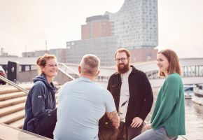 Vier Personen sitzen an der Jan-Fedder-Promenade im Hintergrund sieht man die Elbphilharmonie