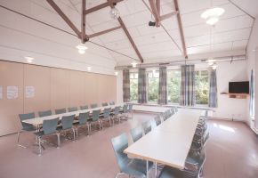 Ein heller Raum mit großen Fenstern, zwei langen Tischen und Stühlen drumherum