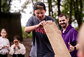 Ein Junge mit Basecap hält ein Holzbrett in seinen Händen. Im Hintergrund sind ein erwachsener Mann und zwei Mädchen zu sehen
