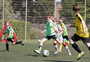 Kinder spielen Fußball auf einem Sportplatz, mehrere Jungen laufen dem Ball nach.