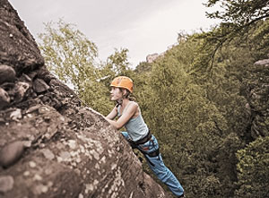 Mädchen mit Helm klettert an einer Felswand im Grünen.